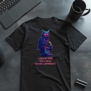 May include: Black t-shirt featuring a pixelated owl with neon blue and pink accents. The owl is perched on a coffee machine, with the text "CAFFEINE DRIVEN DEVELOPMENT" below. A laptop and coffee mug are in the background.