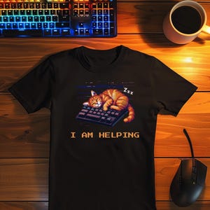 Pode incluir: Camiseta preta com um gato malhado laranja pixelado dormindo em um teclado. O texto "I AM HELPING" está abaixo do gato. Um mouse de computador e uma xícara de café também são visíveis.