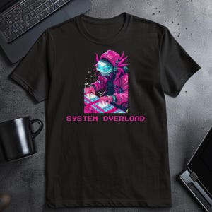 May include: Black t-shirt featuring a vibrant graphic of a figure in a futuristic helmet and pink jacket, operating a keyboard. The text "SYSTEM OVERLOAD" is displayed in pixelated pink letters below the graphic. The shirt is made of a soft material.
