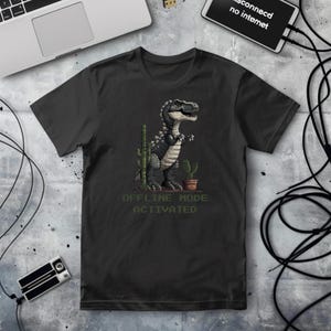 May include: Black t-shirt featuring a pixelated dinosaur illustration with cacti. The text "OFFLINE MODE ACTIVATED" is printed below the graphic. The shirt is displayed on a textured surface with a laptop and other tech accessories.