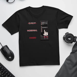 May include: Black t-shirt with a pixelated video game controller design. The design includes the words "EASY", "NORMAL", and "HARD" in red, along with a pixelated hand gesture. A computer mouse and keyboard are visible in the image.