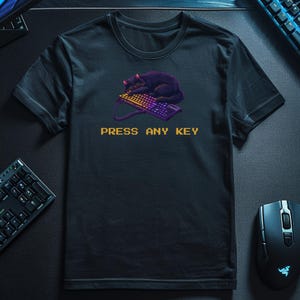 Puede incluir: Camiseta negra con un diseño pixelado de un gato negro durmiendo sobre un teclado. El texto "PRESS ANY KEY" está impreso en amarillo debajo. La camiseta es de manga corta y está hecha de una tela oscura.