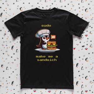 May include: A black short-sleeve t-shirt featuring a graphic of a penguin wearing a chef hat and holding a layered sandwich on a plate. The text above the penguin reads "sudo" and the text below the penguin reads "make me a sandwich" in yellow.