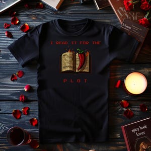 Peut inclure: T-shirt noir avec l'inscription "I READ IT FOR THE PLOT". Le motif représente un livre ouvert avec des flammes et un piment rouge. Le t-shirt est entouré de pétales de rose et d'une bougie.