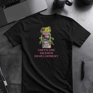 Puede incluir: Camiseta negra con un dibujo de una rana verde con ojos rojos sentada sobre una lata. El texto "CAFFEINE DRIVEN DEVELOPMENT" está impreso en rosa debajo.