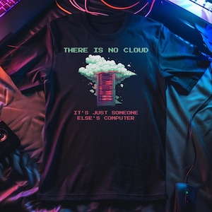 Pode incluir: Uma t-shirt preta de manga curta com um gráfico de um rack de servidores rodeado por uma nuvem verde clara. O texto THERE IS NO CLOUD aparece acima e IT'S JUST SOMEONE ELSE'S COMPUTER em fonte vermelha pixelizada abaixo do rack.
