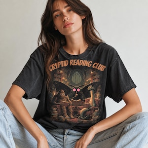 Può includere: Una t-shirt a maniche corte grigio antracite con una stampa grafica che riporta il testo "CRYPTID READING CLUB" in lettere arancioni. L'illustrazione raffigura Bigfoot, una creatura simile a una falena e un serpente marino che leggono.