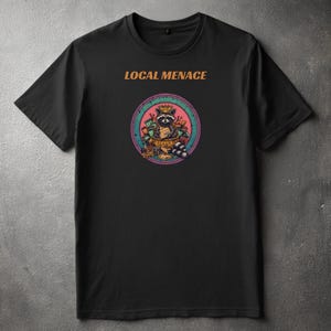 Puede incluir: Camiseta negra con el texto "LOCAL MENACE" sobre un gráfico circular. El gráfico presenta un mapache con una corona, rodeado de dinero y otros objetos. El gráfico tiene acentos rosas, verdes y naranjas.
