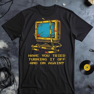 Op de afbeelding: Een zwart T-shirt met korte mouwen en een afbeelding van een gele vintage televisie omwikkeld met geel waarschuwingslint. Onder de televisie staat de tekst: HAVE YOU TRIED TURNING IT OFF AND ON AGAIN? Het ontwerp is in het midden geplaatst.