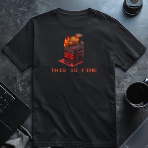 Può includere: T-shirt nero con un cartone animato di un capibara su un rack di server in fiamme, che tiene una tazza. Il testo "THIS IS FINE" è stampato in rosso. Un design umoristico.