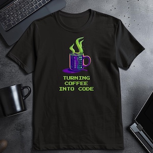 Puede incluir: Camiseta negra con un diseño de taza de café morada, código binario y vapor verde. El texto "TURNING COFFEE INTO CODE" está en verde. Un portátil y una taza negra están en el fondo.