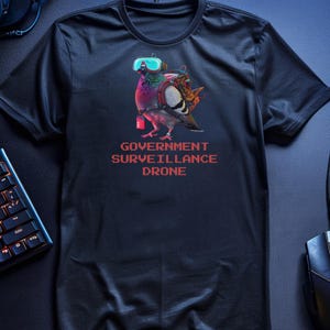 以下が含まれることがあります： 黒いTシャツ。ゴーグルとバックパックをつけたカラフルなハトのグラフィックが特徴で、赤字で「GOVERNMENT SURVEILLANCE DRONE」と書かれています。デザインはTシャツの中央に配置されています。