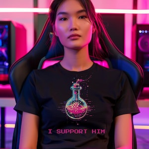 Könnte beinhalten: Schwarzes T-Shirt mit einer farbenfrohen Grafik einer Zaubertrankflasche mit rosa Flüssigkeit und Blasen. Der Text "I SUPPORT HIM" ist darunter gedruckt. Die Person sitzt in einem schwarzen Gaming-Stuhl.