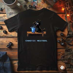 May include: Black t-shirt featuring a pixelated black cat spilling water from a blue glass. The cat sits on a wooden surface, with the words "CHAOTIC NEUTRAL" in white below. The shirt is displayed on a wooden surface with dice.