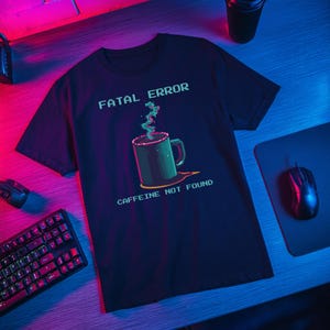 Puede incluir: Camiseta azul marino oscuro con un gráfico pixelado de una taza de café y el texto "FATAL ERROR CAFFEINE NOT FOUND" en verde y blanco. La taza tiene un asa verde y emite vapor verde.