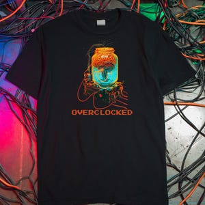 Op de afbeelding: Zwart t-shirt met een afbeelding van een hersen in een pot, bovenop een computercomponent, met het woord "OVERCLOCKED" in oranje. De hersenen zijn oranje en rood, de pot is blauw en de component is geel en groen.