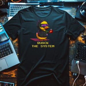Op de afbeelding: Zwart t-shirt met een cartoon eend met een hoodie en zonnebril, die een klein rood object vasthoudt. De tekst "QUACK THE SYSTEM" staat in gele pixelletter. Het ontwerp suggereert een thema van technologie of hacking.