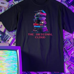 May include: Black t-shirt with a graphic of a stack of colourful VHS tapes, and the pink text "THE ORIGINAL CLOUD". The design has a retro feel, ideal for fans of vintage media and nostalgic items.