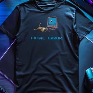 May include: Navy blue t-shirt featuring a pixelated illustration of a dead rodent next to a retro computer monitor. The text "FATAL ERROR" is printed in blue block letters below the graphic. The shirt is made of a soft material.