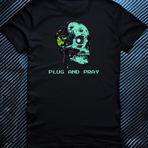 May include: Black t-shirt with a teal skull graphic, incorporating mechanical elements and the text "PLUG AND PRAY". The skull design includes a glowing eye and is detailed with purple, green, and yellow accents in a pixelated font.