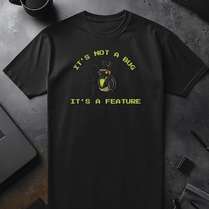 Può includere: T-shirt nera con la scritta "IT'S NOT A BUG, IT'S A FEATURE" in giallo. Al centro c'è un gufo dei cartoni animati con cilindro e bastone. La maglietta è realizzata in materiale morbido.