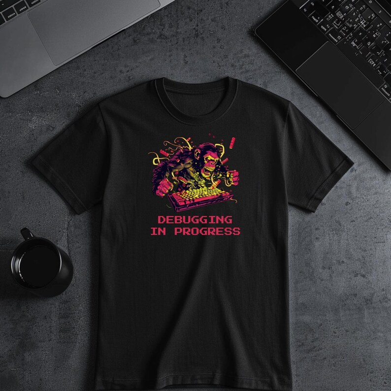 May include: Black t-shirt with a vibrant graphic of a gorilla working on a keyboard. The design uses pink, yellow, and green hues, and the text "DEBUGGING IN PROGRESS" is displayed below the image.