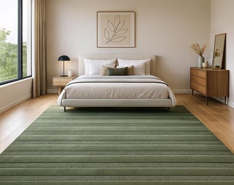 Puede incluir: Una alfombra verde con rayas horizontales en diferentes tonos de verde, colocada en un dormitorio. La alfombra es el punto focal, con una cama, una mesita de noche y una cómoda en el fondo. La habitación tiene una paleta de colores neutros con luz natural.
