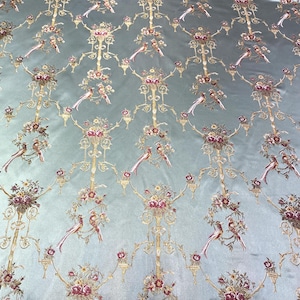 May include: A light blue fabric with an ornate pattern featuring gold filigree, floral bouquets, and birds. The design is repeated across the fabric, creating a luxurious and detailed textile. The fabric has a subtle sheen, enhancing the intricate details.