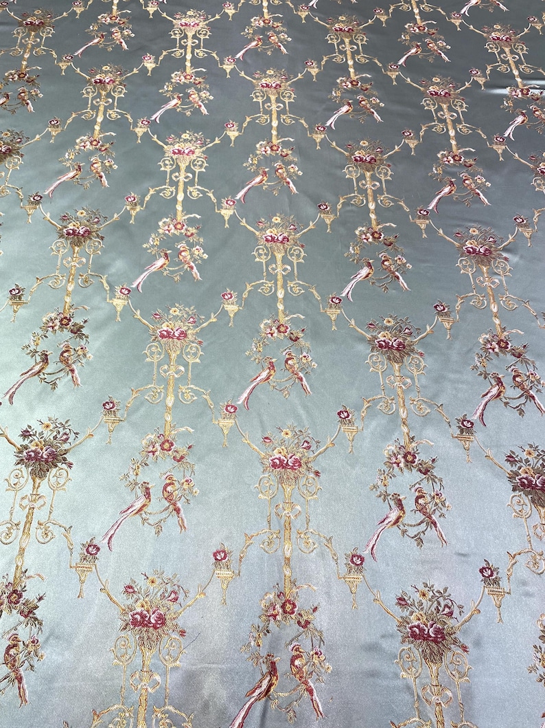 May include: A light blue fabric with an ornate pattern featuring gold filigree, floral bouquets, and birds. The design is repeated across the fabric, creating a luxurious and detailed textile. The fabric has a subtle sheen, enhancing the intricate details.