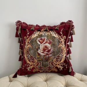 May include: A decorative burgundy pillow with a floral rose design. The pillow features a central oval image of roses in shades of red and cream, framed by ornate gold detailing. Tassels adorn the edges, adding a touch of elegance.