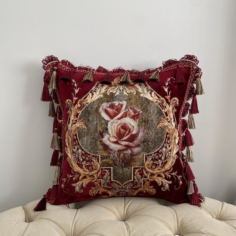 May include: A decorative burgundy pillow with a floral rose design. The pillow features a central oval image of roses in shades of red and cream, framed by ornate gold detailing. Tassels adorn the edges, adding a touch of elegance.
