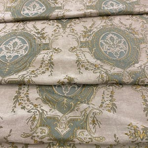 May include: Close-up of folded fabric with a repeating pattern. The design features ornate, oval medallions in light blue and gold, set against a neutral background. Floral and scroll details fill the spaces between the medallions.