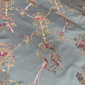 May include: A close-up of a fabric with a repeating pattern of birds, flowers, and ornate gold-colored candelabra-like designs on a pale green background. The birds have red, pink, and white feathers. The flowers are red and pink.
