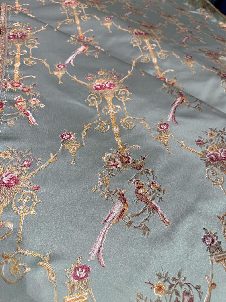 May include: A close-up of a fabric with a repeating pattern of birds, flowers, and ornate gold-colored candelabra-like designs on a pale green background. The birds have red, pink, and white feathers. The flowers are red and pink.