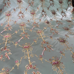 May include: A light blue fabric with an ornate pattern of gold filigree, flowers, and birds. The design is repeated across the surface, creating a luxurious and detailed textile. The birds are depicted in shades of red, brown, and white.