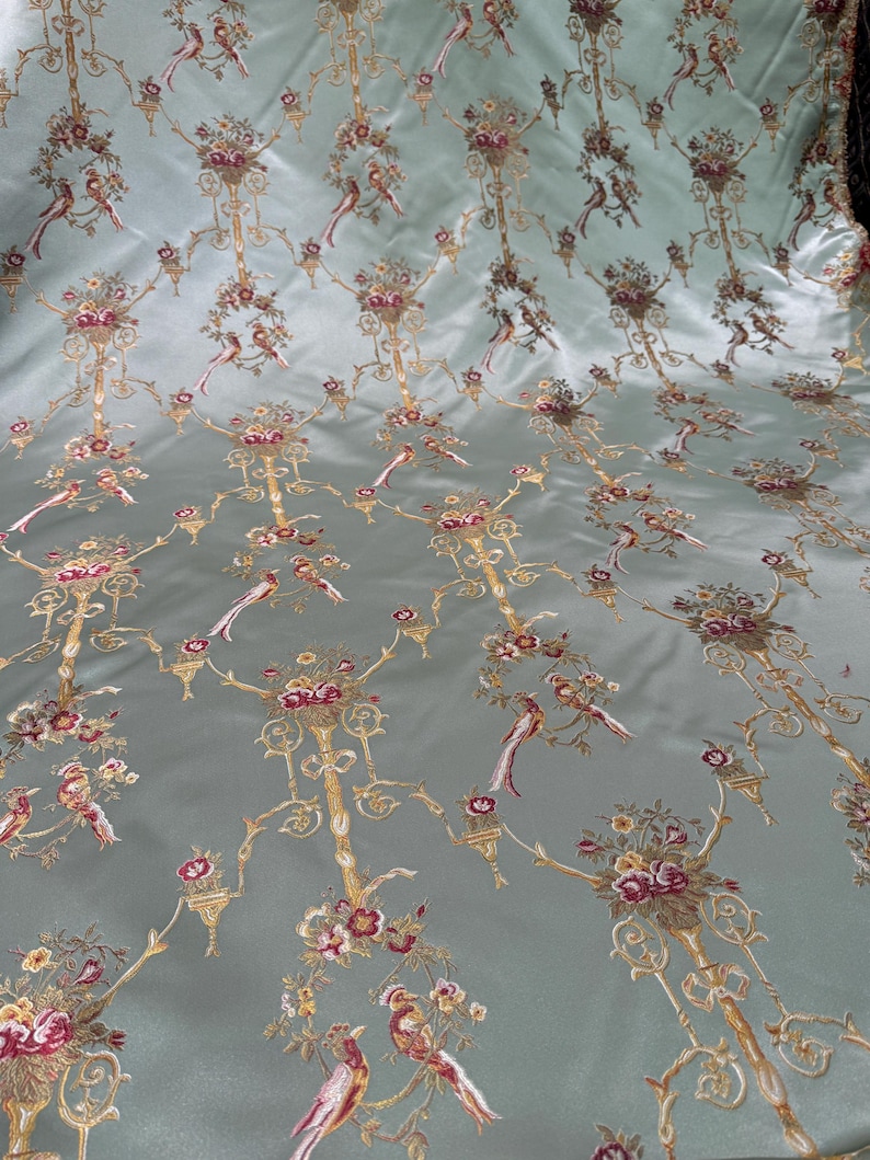 May include: A light blue fabric with an ornate pattern of gold filigree, flowers, and birds. The design is repeated across the surface, creating a luxurious and detailed textile. The birds are depicted in shades of red, brown, and white.