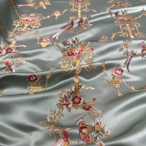 May include: Silky, sage green fabric with an embroidered pattern of birds, floral arrangements, and ornate gold-colored architectural elements. The fabric has a subtle sheen, suggesting a luxurious material suitable for apparel or home decor.