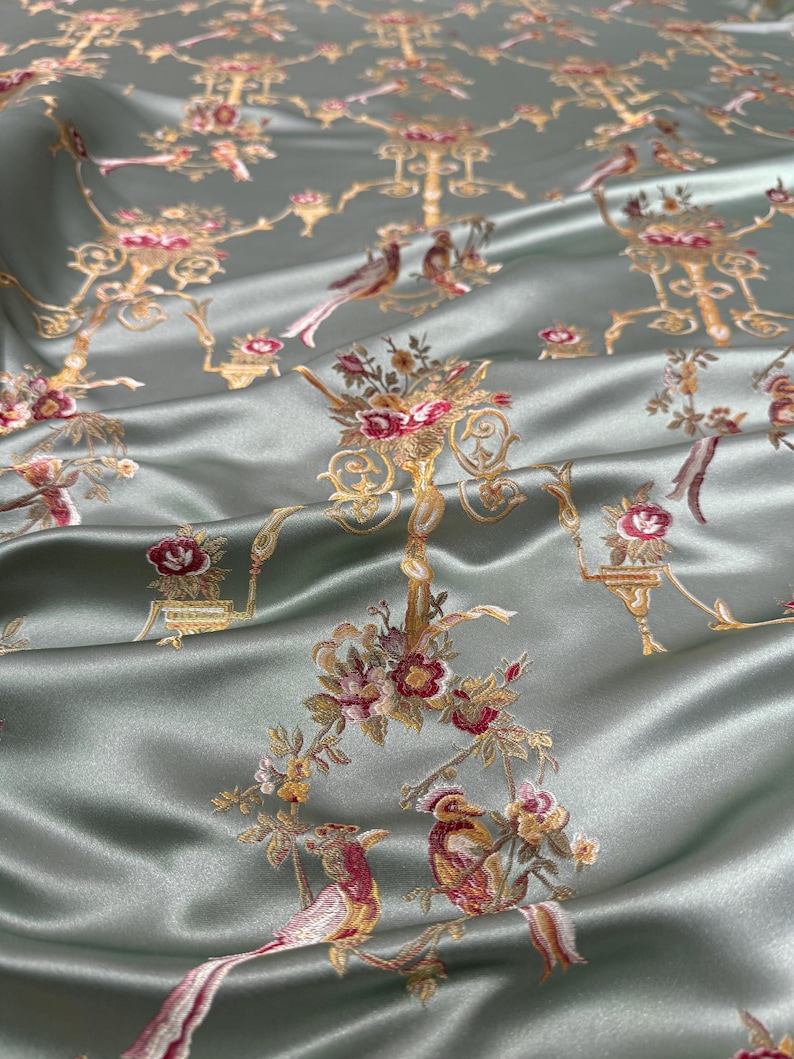 May include: Silky, sage green fabric with an embroidered pattern of birds, floral arrangements, and ornate gold-colored architectural elements. The fabric has a subtle sheen, suggesting a luxurious material suitable for apparel or home decor.