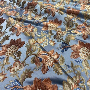 Op de afbeelding: Close-up van een stof met een bloemenpatroon. De stof heeft een lichtblauwe achtergrond met grote, gedetailleerde bloemen in tinten bruin, goud en blauw. De stof heeft een lichte glans, wat wijst op satijn of zijde.