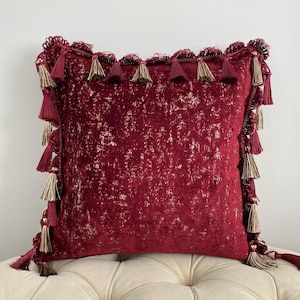May include: A square decorative pillow in a deep red hue with gold accents. The pillow is trimmed with a fringe of tassels in matching red and gold tones, adding a touch of elegance to the design. The pillow is placed on a cream-colored tufted surface.