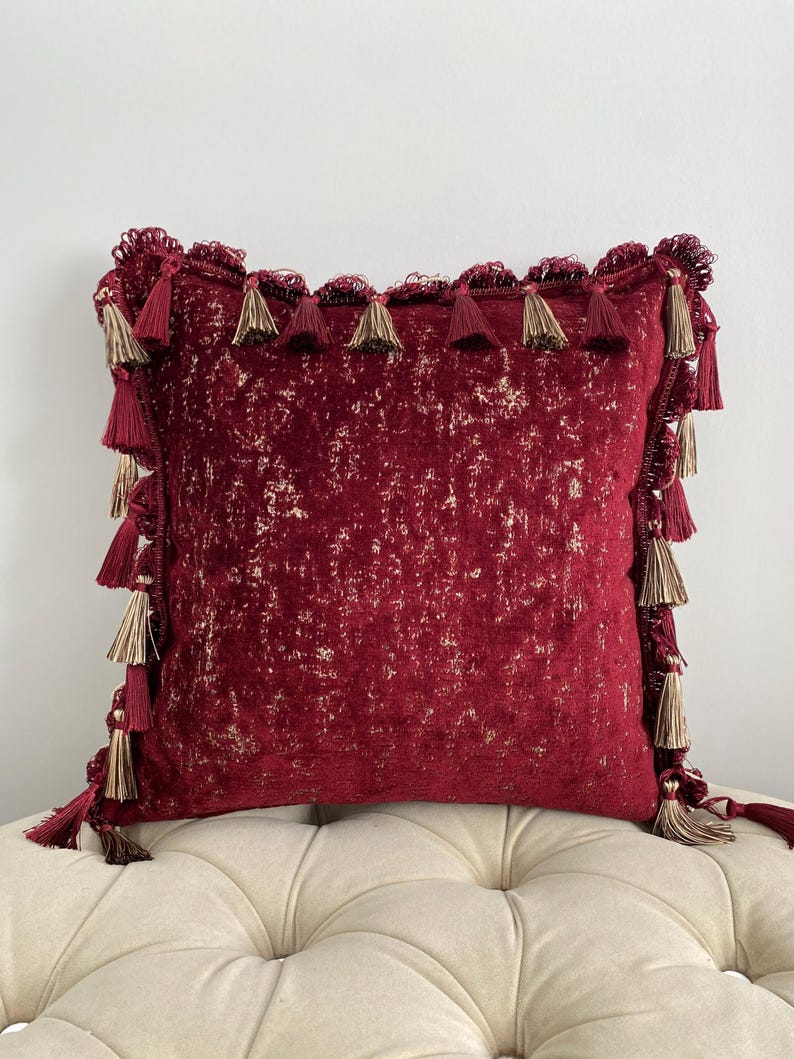 May include: A square decorative pillow in a deep red hue with gold accents. The pillow is trimmed with a fringe of tassels in matching red and gold tones, adding a touch of elegance to the design. The pillow is placed on a cream-colored tufted surface.