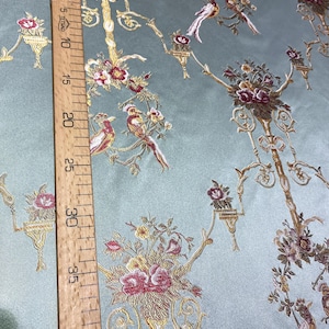 May include: A light blue fabric with an embroidered floral and bird design. The design includes gold-colored branches, red and pink flowers, and detailed birds. A wooden ruler shows measurements up to 40 inches.