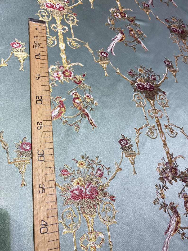 May include: A light blue fabric with an embroidered floral and bird design. The design includes gold-colored branches, red and pink flowers, and detailed birds. A wooden ruler shows measurements up to 40 inches.