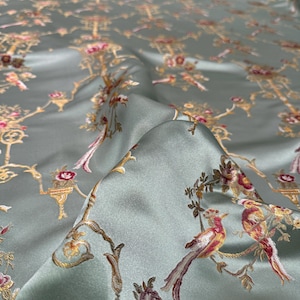 May include: A close-up of a folded, silky fabric with a repeating pattern of birds, flowers, and ornate architectural elements in gold, pink, and red on a pale green background. The fabric has a slight sheen.