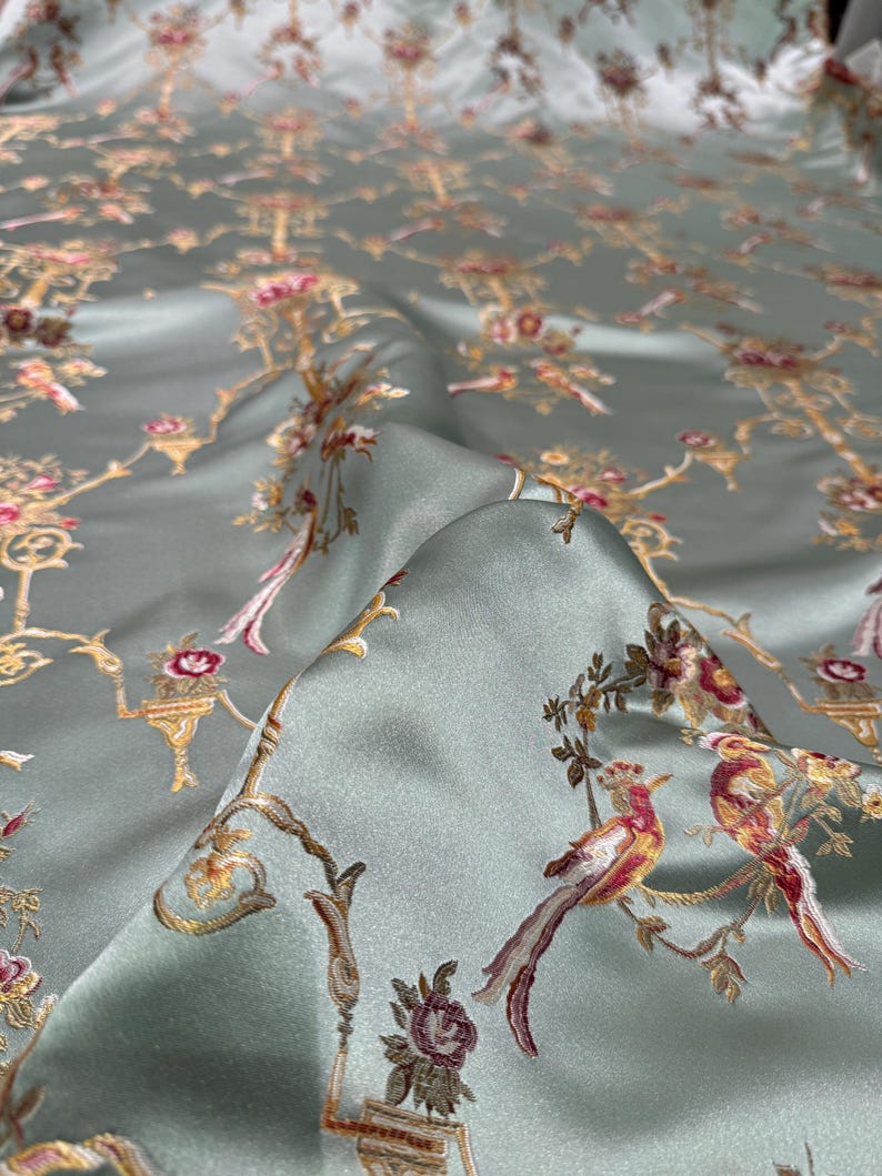 May include: A close-up of a folded, silky fabric with a repeating pattern of birds, flowers, and ornate architectural elements in gold, pink, and red on a pale green background. The fabric has a slight sheen.