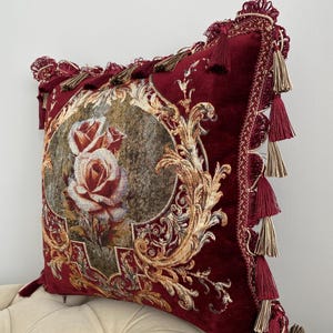 May include: A burgundy decorative pillow with a floral rose design. The pillow features gold filigree and tassels along the edges. The pillow is approximately 18 inches square.
