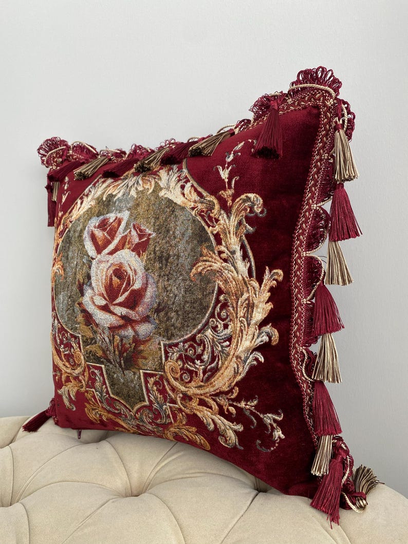 May include: A burgundy decorative pillow with a floral rose design. The pillow features gold filigree and tassels along the edges. The pillow is approximately 18 inches square.