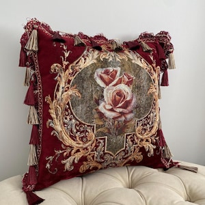 May include: A decorative burgundy pillow with a floral design. The pillow features a central image of roses within an ornate frame. Gold and beige accents complement the burgundy fabric. Tassels adorn the edges, adding a touch of elegance to this home decor item.