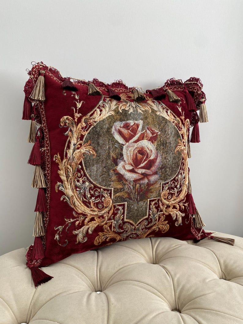 May include: A decorative burgundy pillow with a floral design. The pillow features a central image of roses within an ornate frame. Gold and beige accents complement the burgundy fabric. Tassels adorn the edges, adding a touch of elegance to this home decor item.