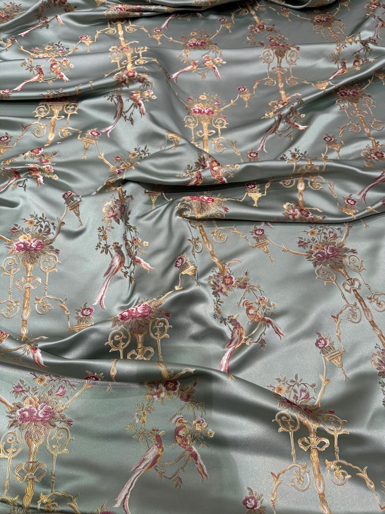 May include: Silky, sage green fabric with a repeating pattern of ornate gold candelabras, birds, and floral elements in shades of pink and burgundy. The fabric has a subtle sheen, suggesting a luxurious material suitable for apparel or home decor.